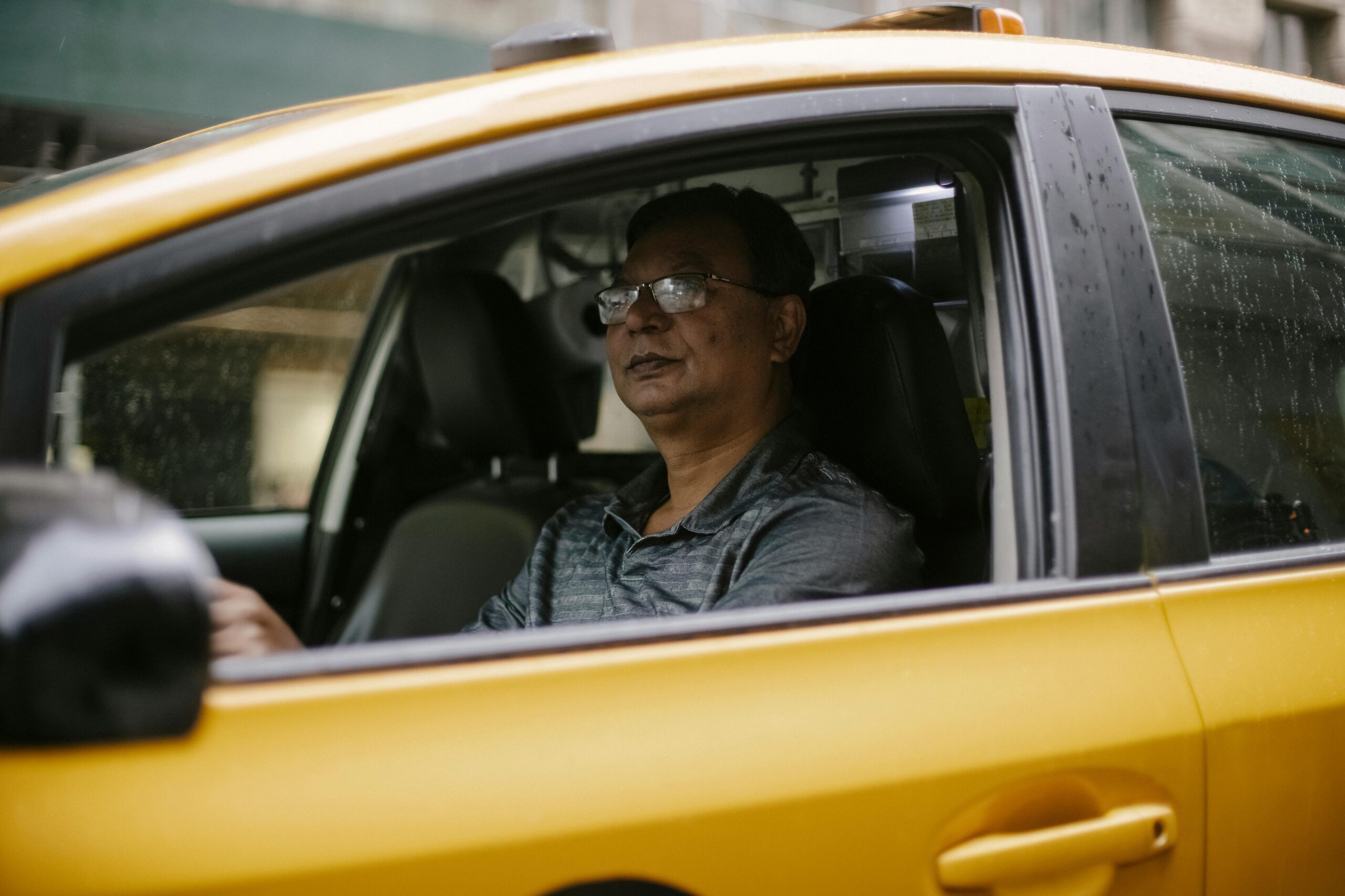 Middle-aged man driving a yellow taxi through a rainy cityscape.