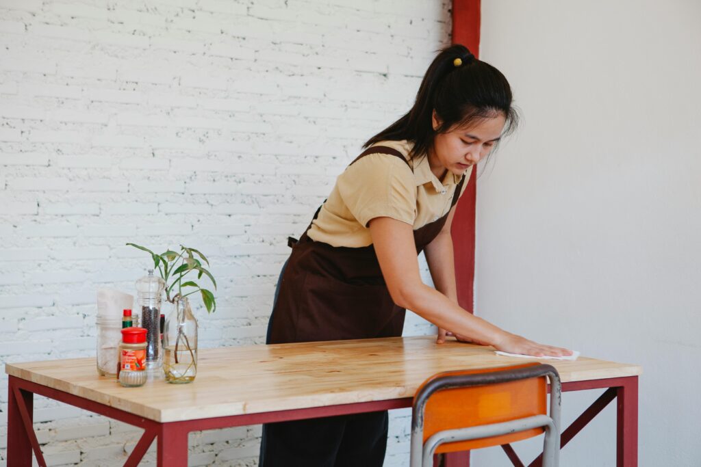Asian woman wiping a wooden table in an indoor setting with cleaning items.